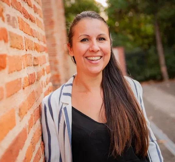 Femme souriante, elle a son corps appuyer sur un mur de brique rouge très présent dans la ville de Toulouse.